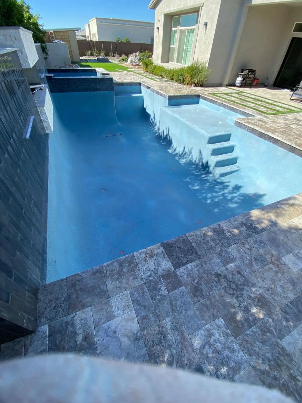 Long blue pool with a stone wall water feature and a gray travertine deck