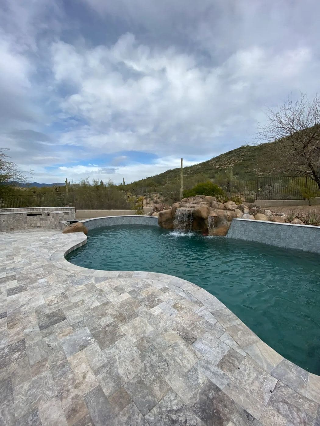 Curved pool with a rock waterfall and travertine deck below a desert mountain