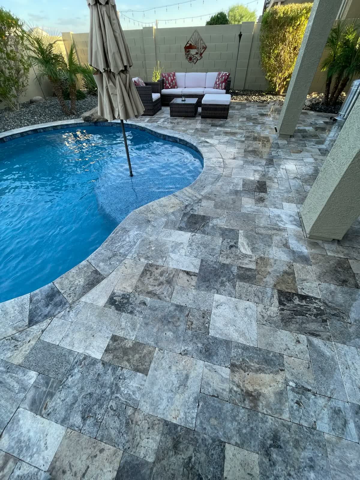 Curved blue pool with a silver travertine deck and patio seating under an umbrella