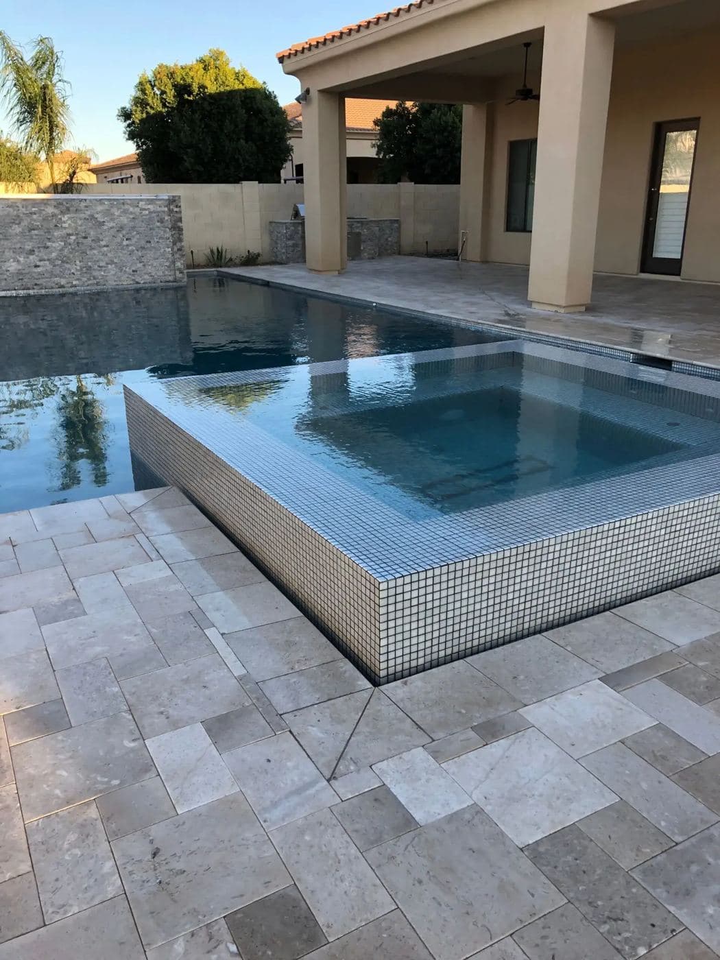 Raised spa wrapped in glass tile next to a pool with a travertine deck
